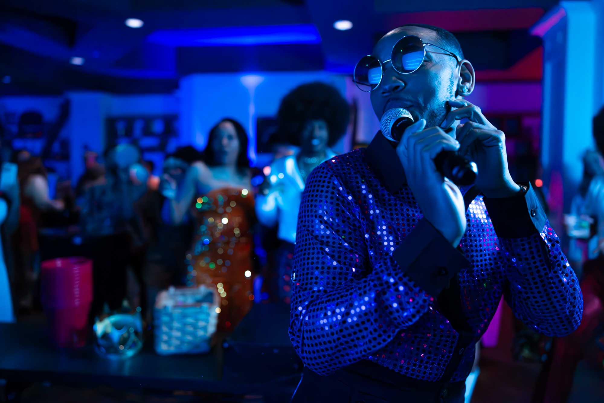 MC in a sequined jacket and round shades holding a microphone at a themed Denver corporate party under blue stage lighting.