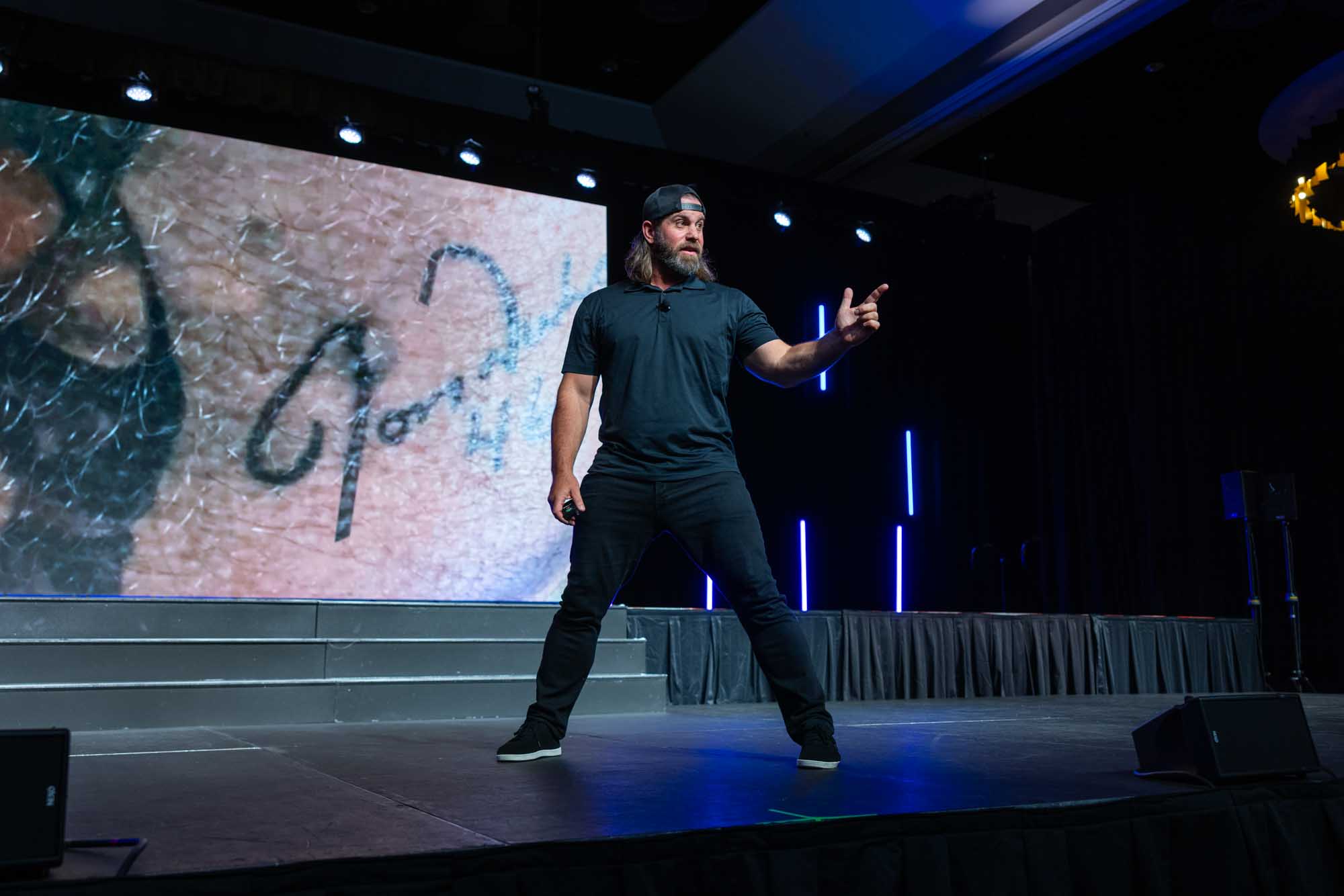 Keynote speaker on stage at a Denver corporate conference, gesturing in front of a large LED presentation screen.