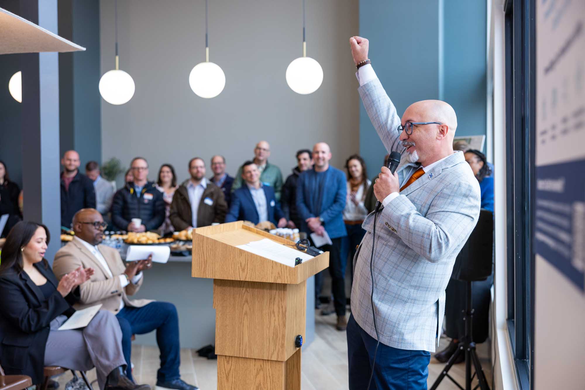 Corporate event speaker with a raised fist rallying staff at a Denver in-office gathering, pendant globe lights overhead.