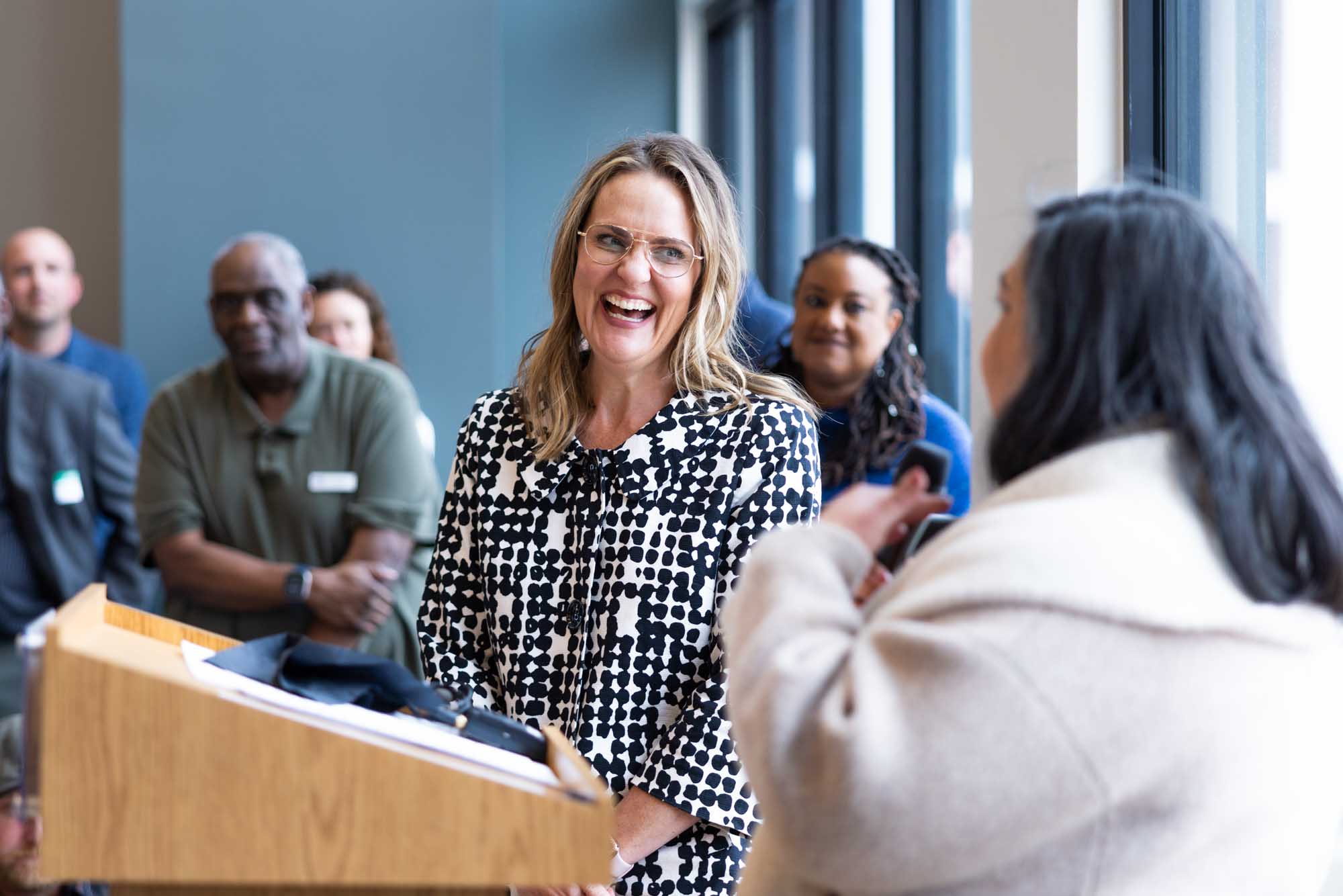 Corporate event speaker laughing at the podium during an in-office Denver company gathering, coworkers listening behind her.