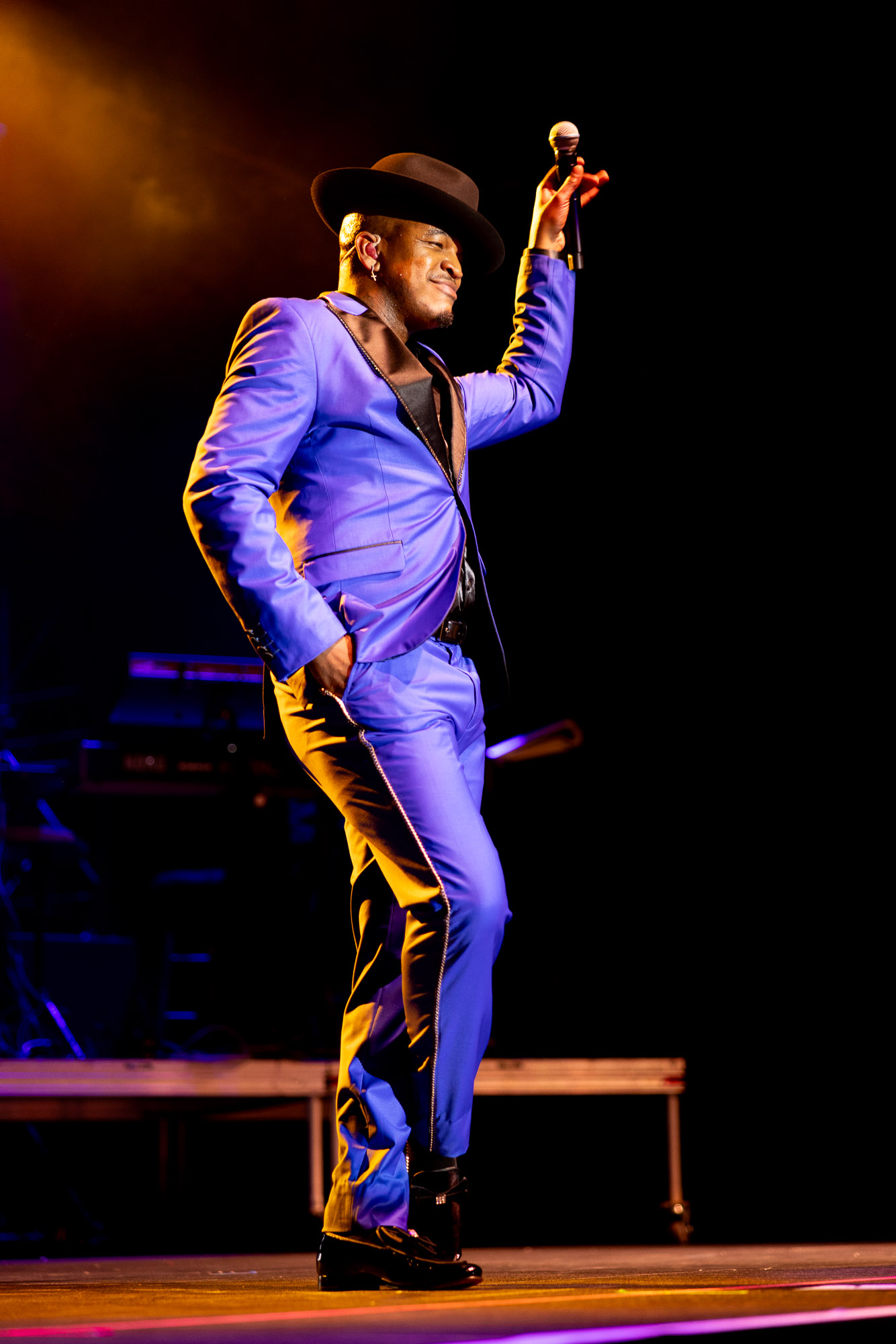 Live music performer in a purple suit and brimmed hat on a theatrically lit Denver concert stage, microphone raised.