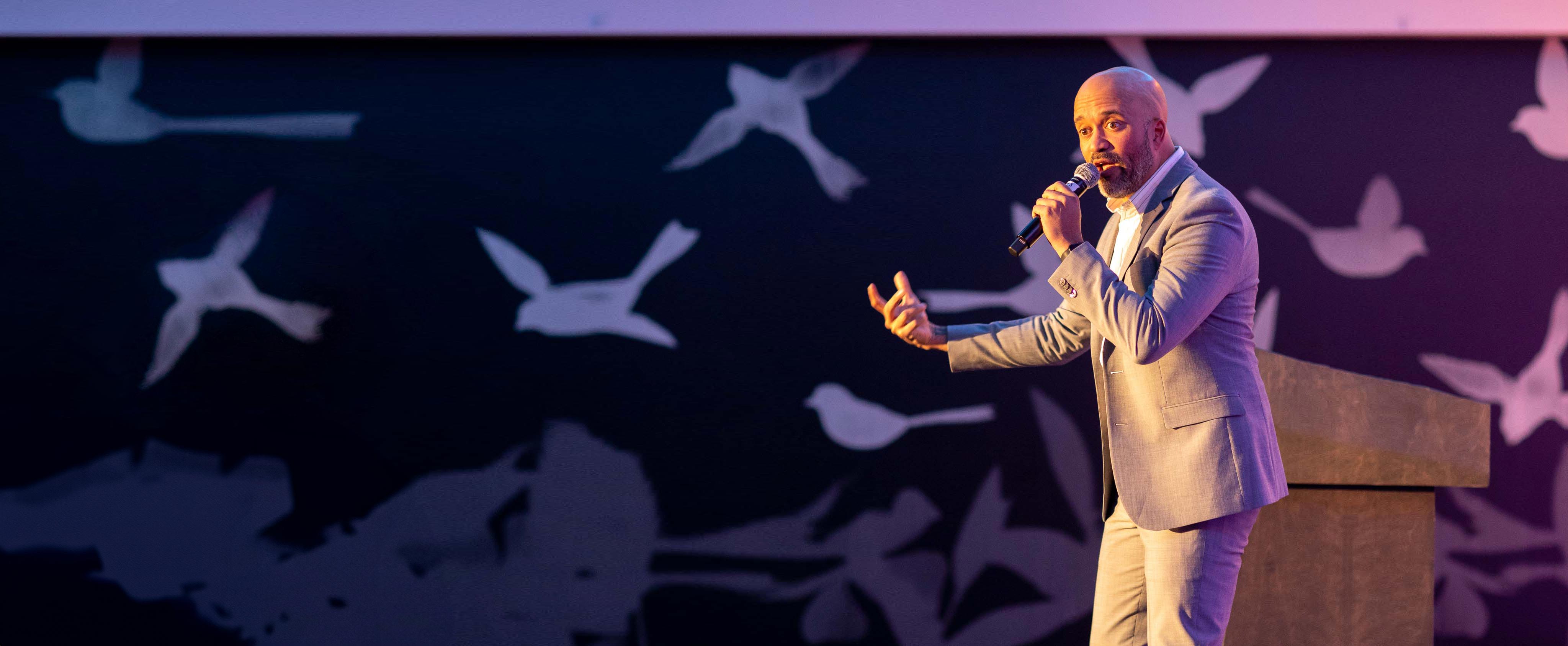 Corporate keynote speaker in a grey suit gesturing mid-speech at a Denver conference, bird-motif stage backdrop behind him.