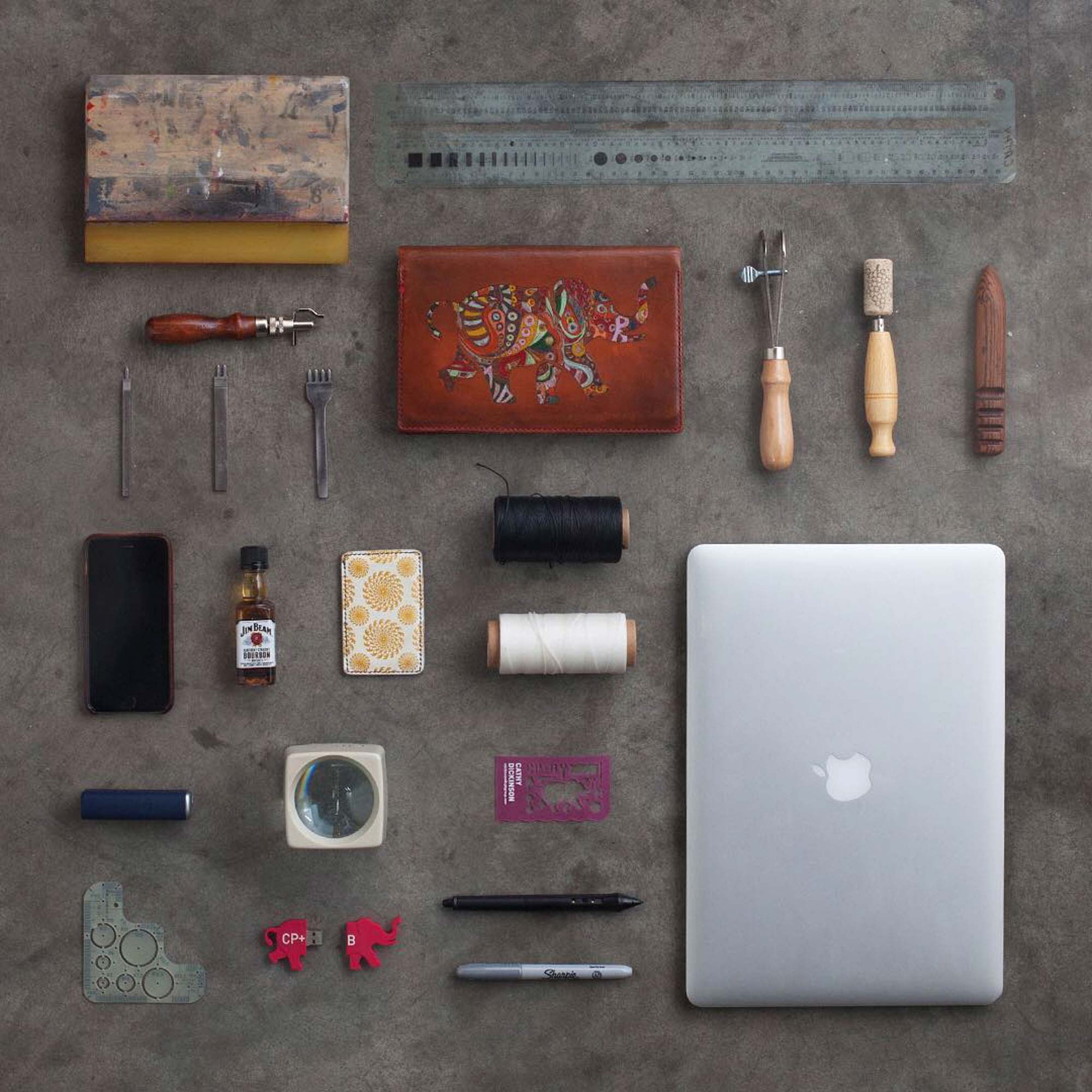 Knolling-style still life of items from a CP+B art-director's desk — leather tools, a MacBook, thread spools, a painted leather wallet and other small desk objects arranged on weathered concrete.