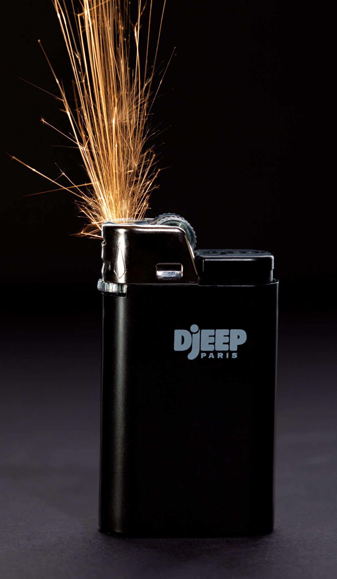 Studio product photograph of a Djeep Paris lighter on a black background with gold sparks arcing from its flint wheel.