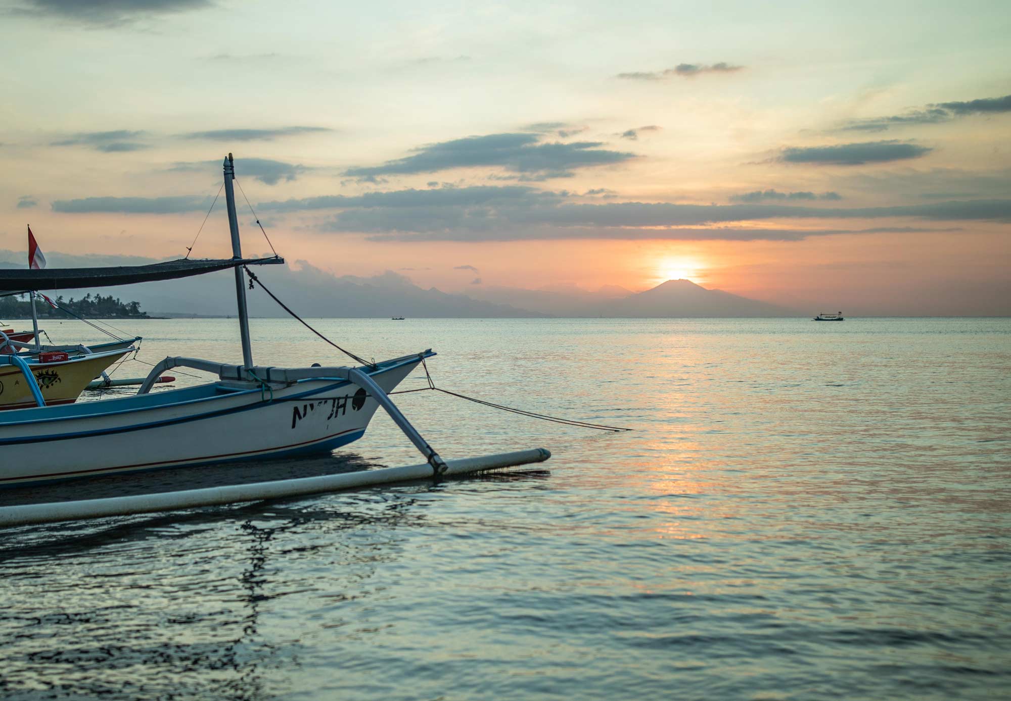 Sunrise over the Bali Sea from Lovina Beach, with a traditional Balinese jukung outrigger in the foreground and a volcano silhouetted on the horizon.