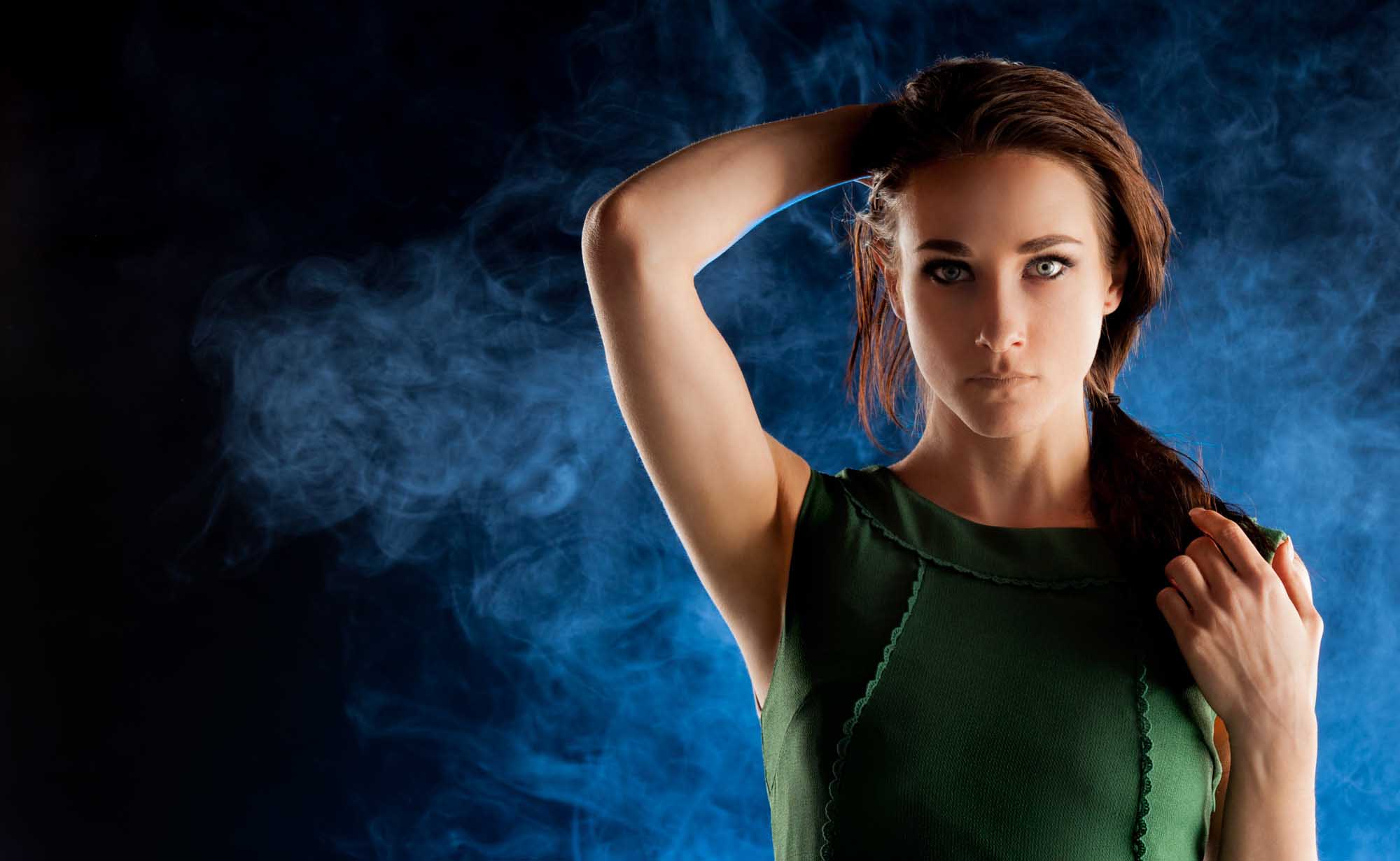 Moody fashion portrait of a woman in a dark green dress against a cloud of blue-lit smoke, with dramatic side-light.