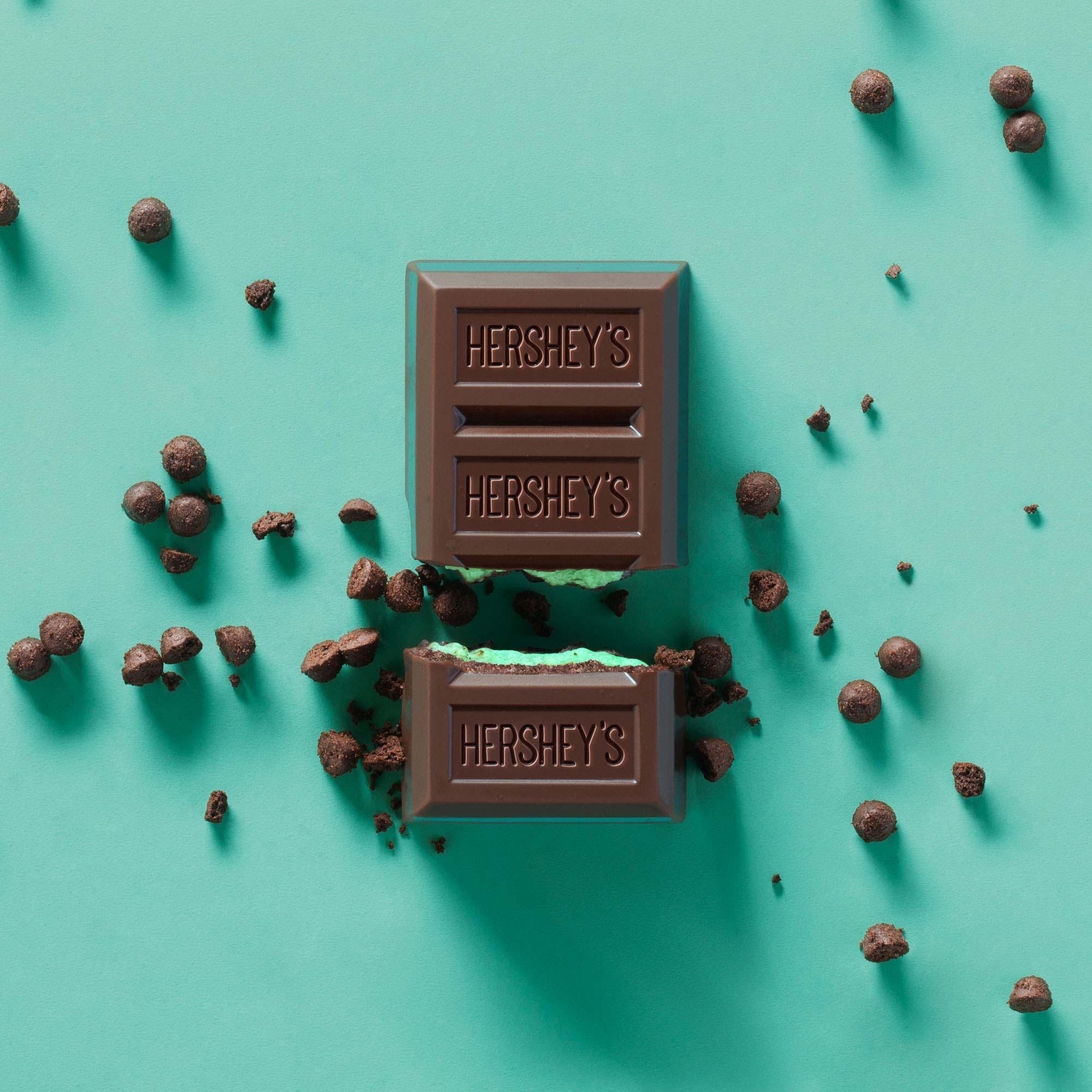 Social-content still life of a Hershey's Cookie Layer Crunch Mint bar broken to reveal its mint filling, with scattered cookie-crunch pieces on a mint-green background.