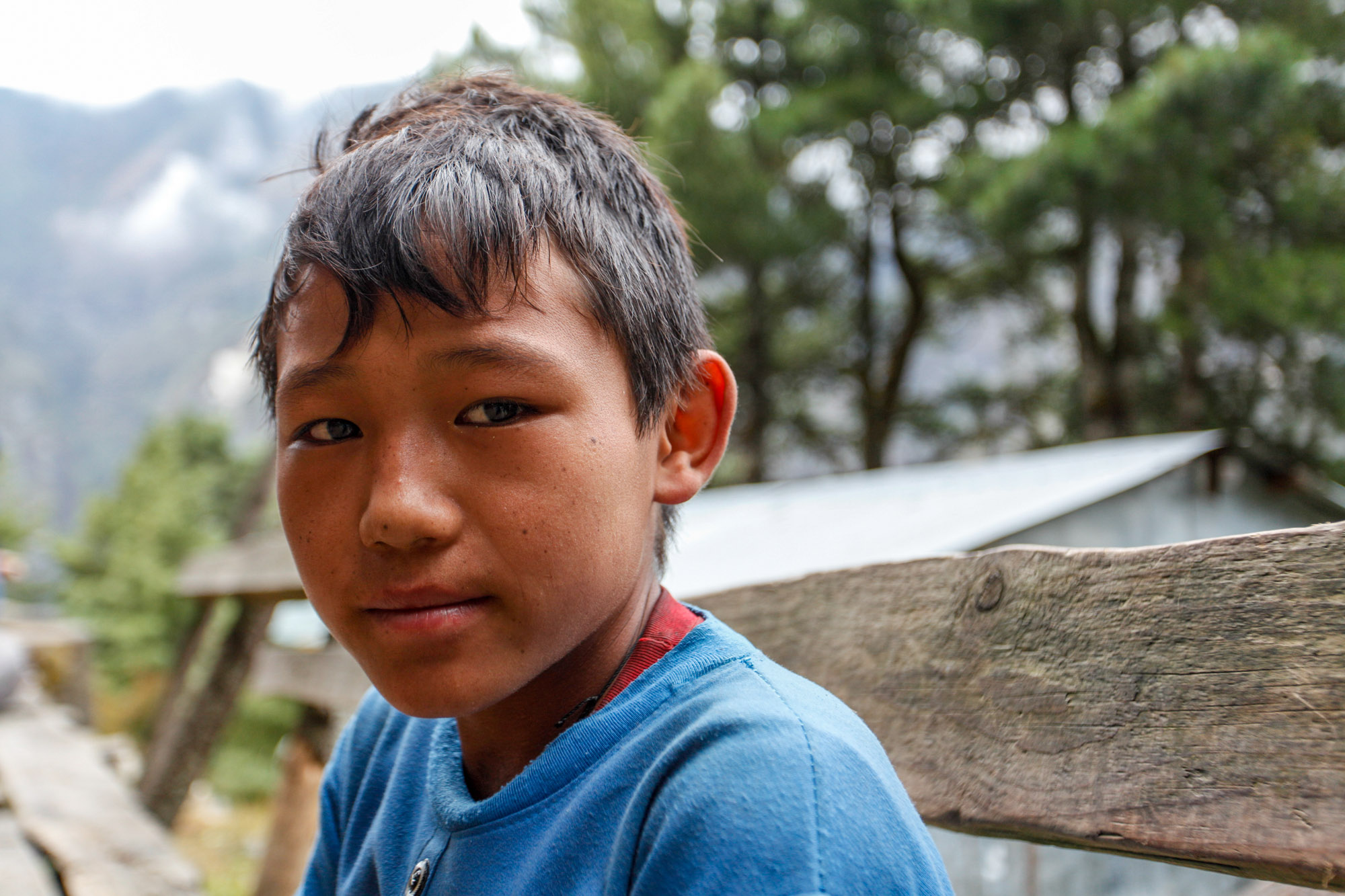 Portrait of a young Nepali child porter in a blue shirt resting against a wooden fence along the Everest Base Camp trail.