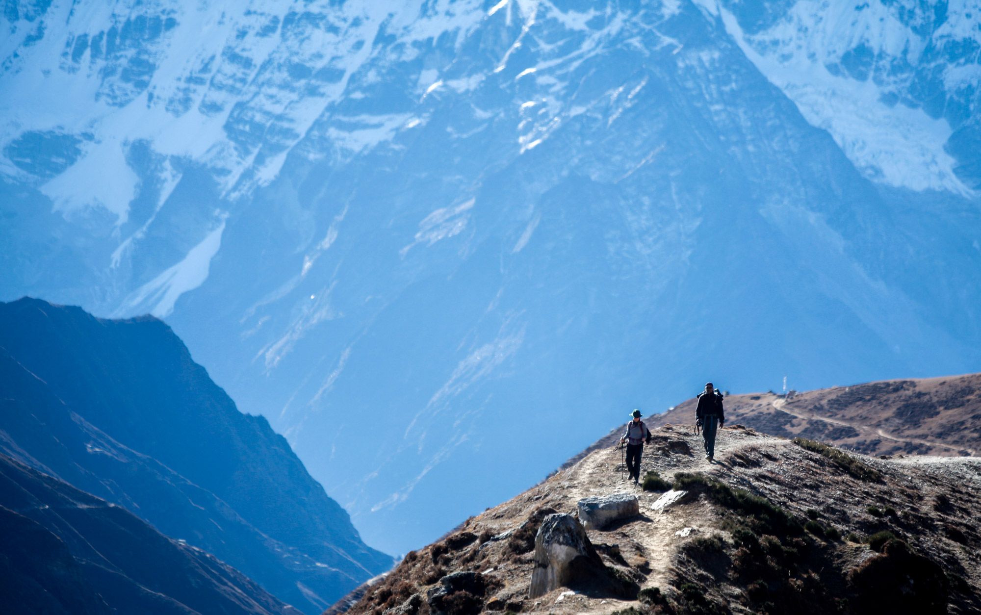 Two trekkers silhouetted on a ridge trail with massive snow-dusted Himalayan walls behind them on the Everest Base Camp trek, Nepal.