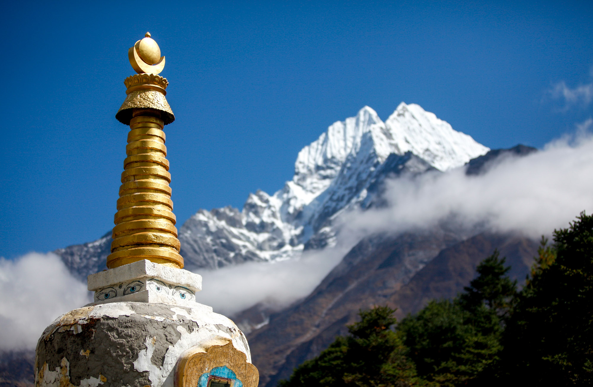 A gold-topped Tibetan Buddhist stupa stands in the foreground with snow-capped Himalayan peaks and drifting clouds behind it — Khumbu region, Nepal.