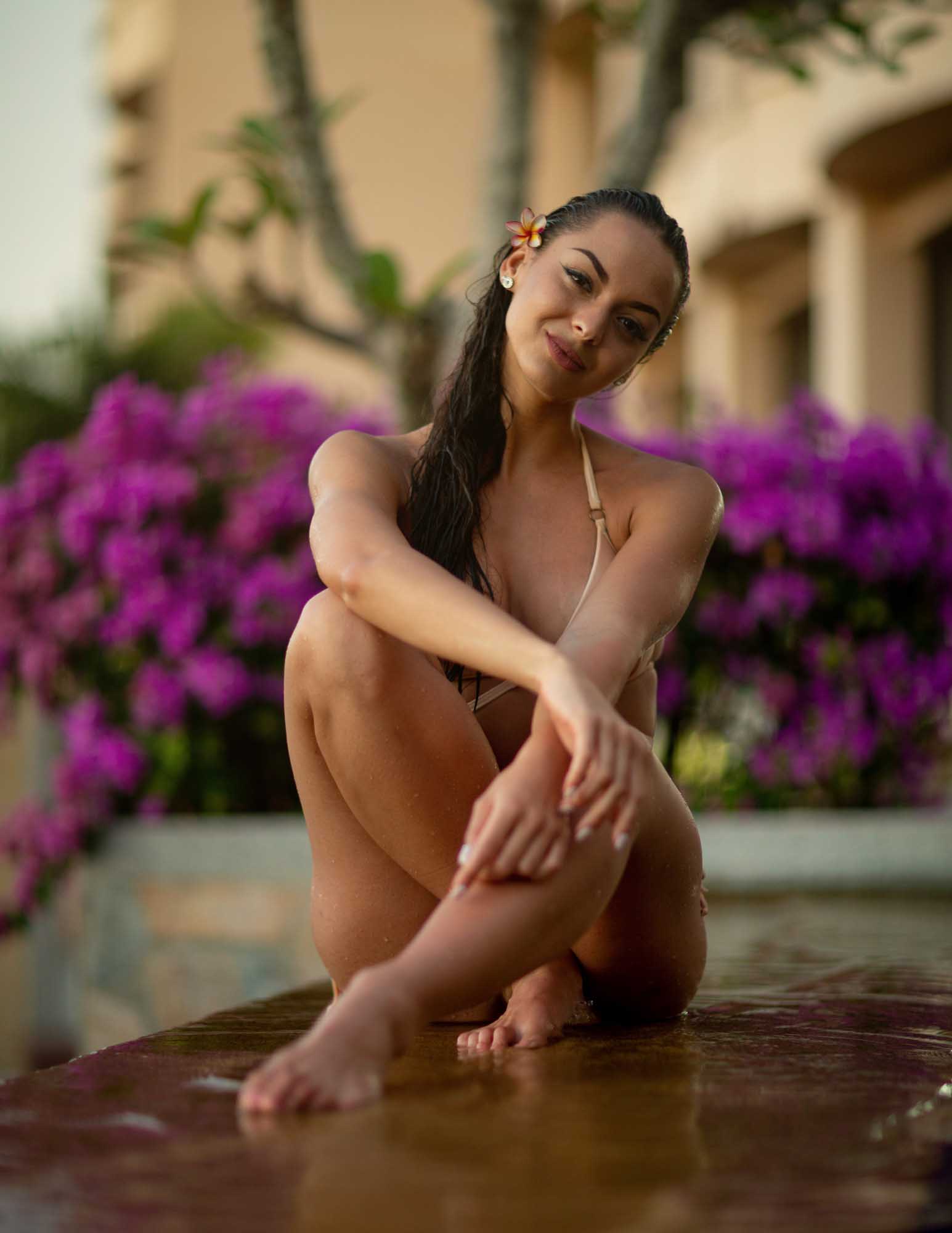 Eniko Kesmarki crouches at the edge of a resort pool beside a burst of magenta bougainvillea, a frangipani tucked behind her ear — Krabi, Thailand.