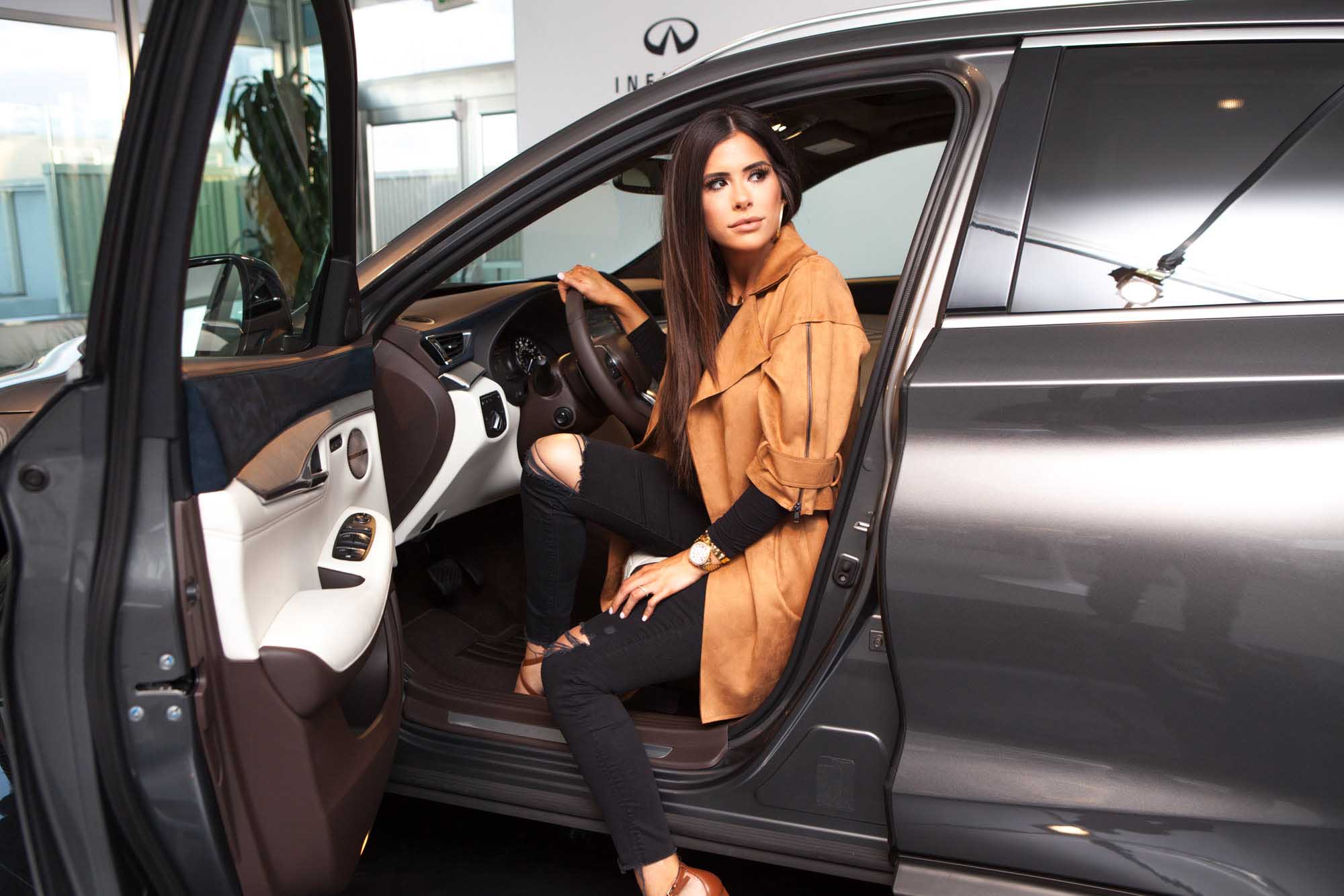 Emily Ann Gemma (@emilyanngemma) sits in the driver's seat of a grey Infiniti QX50 with the door open at an Infiniti showroom, wearing a tan blazer and ripped black jeans.