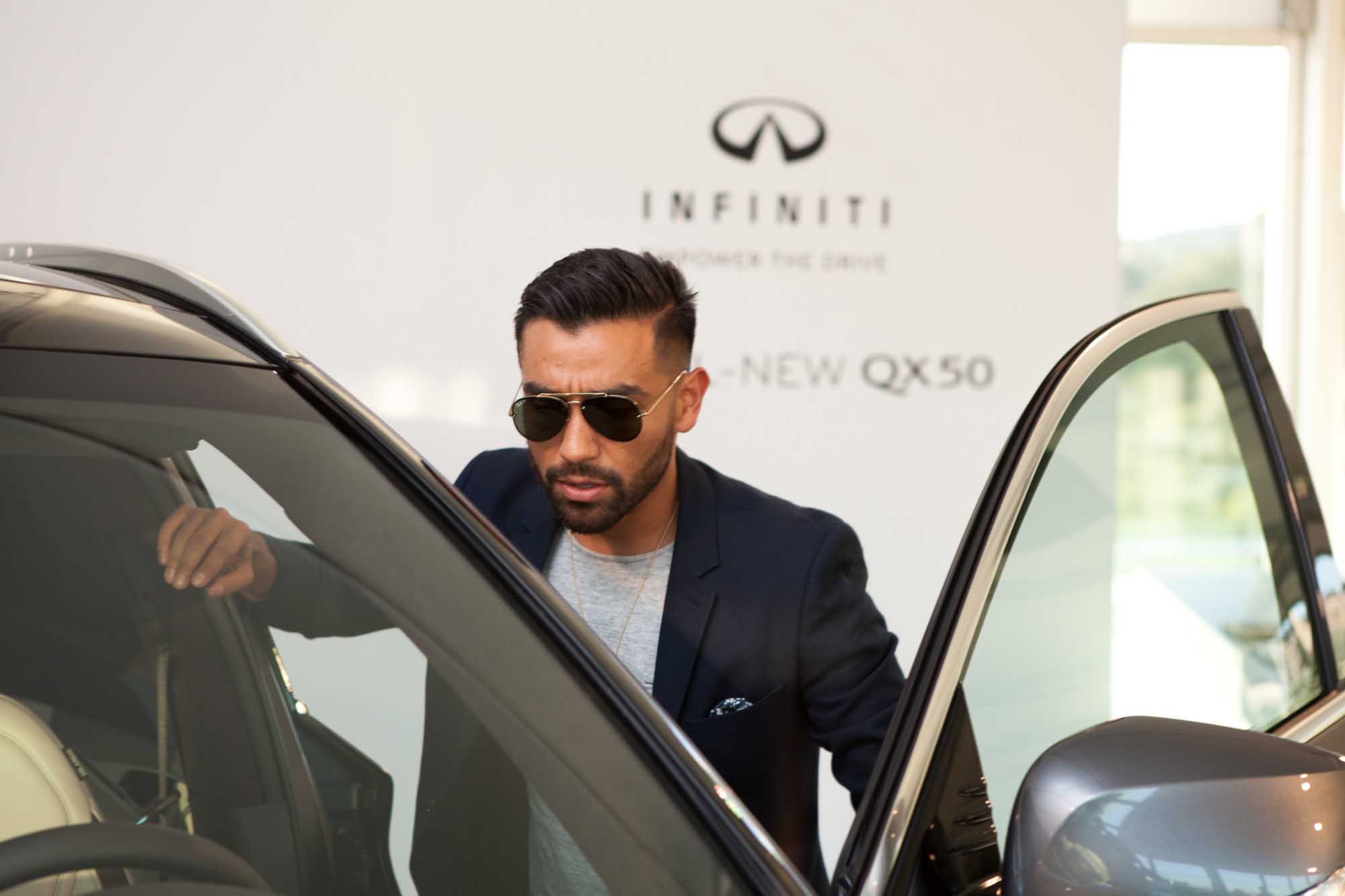 J Fig (@rule_of_thumbs) steps out of an Infiniti QX50 at an Infiniti showroom, wearing a navy blazer and sunglasses.