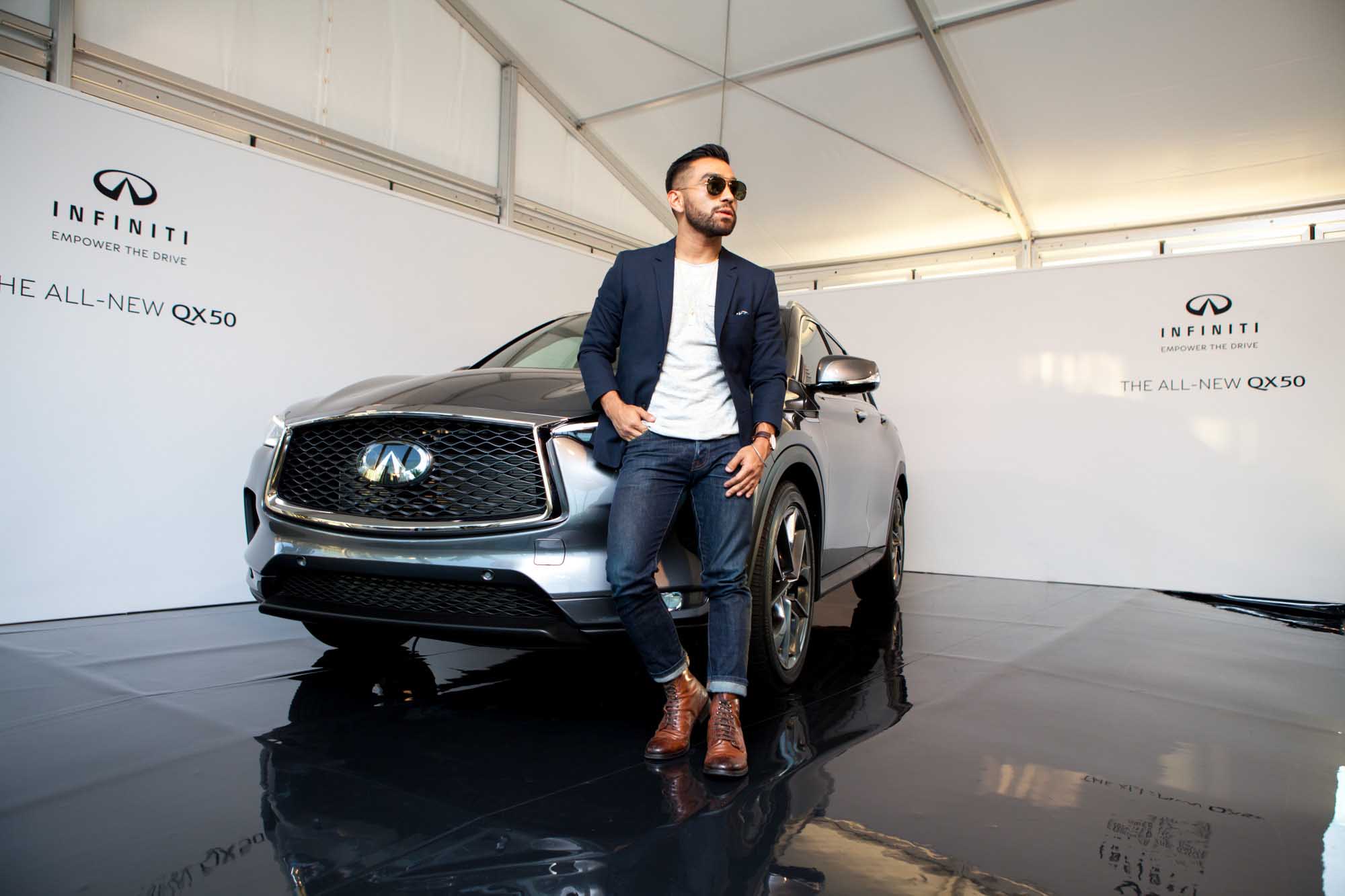 J Fig (@rule_of_thumbs) leans against the front fender of an Infiniti QX50 on the black showroom floor, in a navy blazer, jeans, and brown boots.