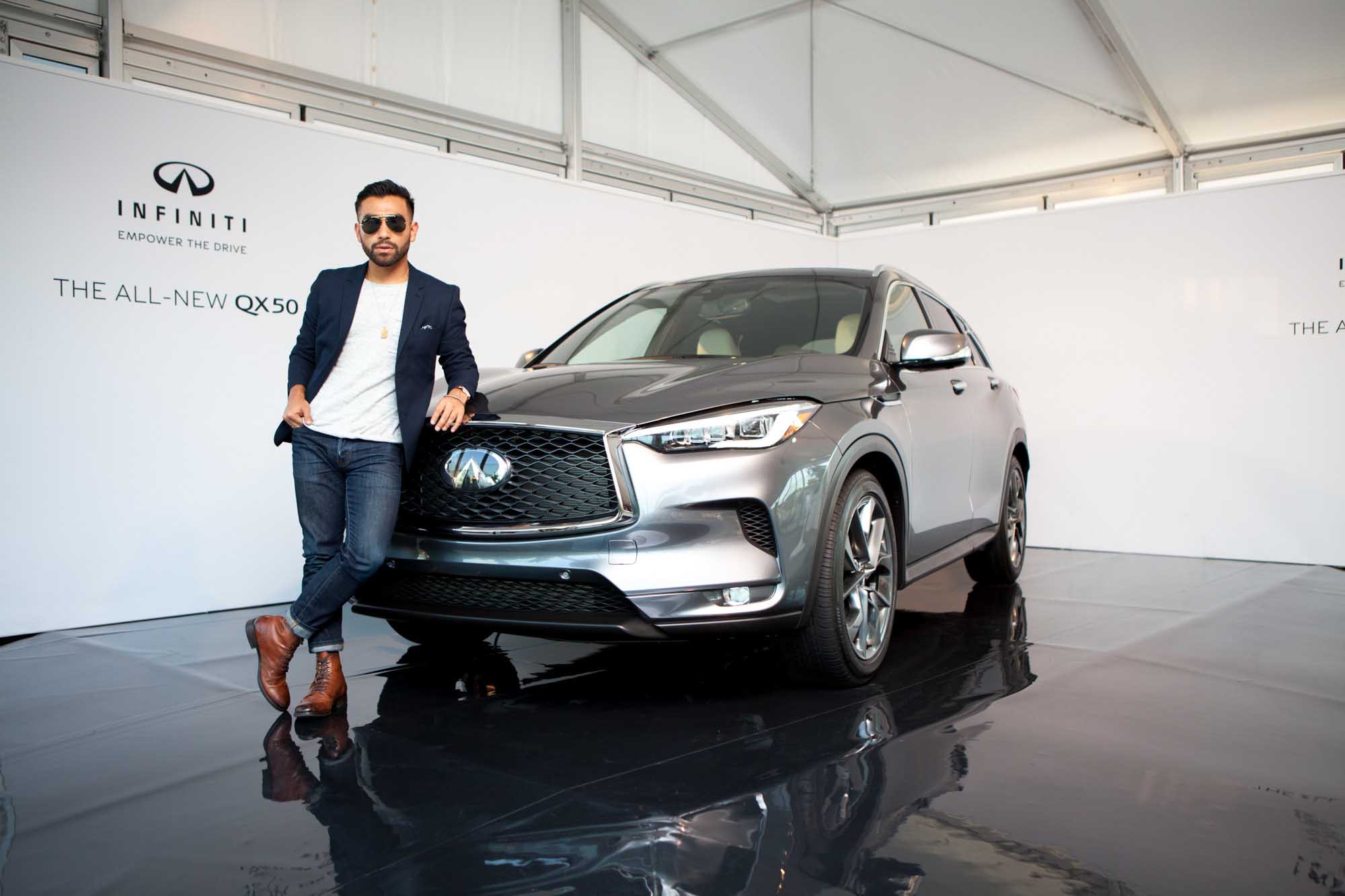 J Fig (@rule_of_thumbs) poses beside the front grille of an Infiniti QX50 in an Infiniti showroom — navy blazer over a white tee, jeans, brown boots.