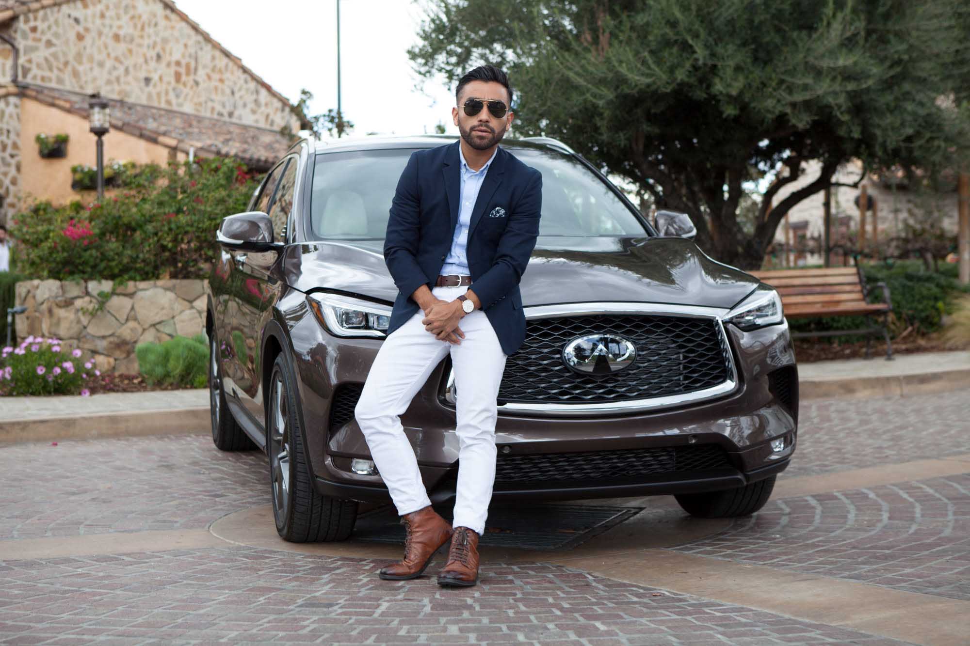 J Fig (@rule_of_thumbs) sits on the hood of an Infiniti QX50 in a navy blazer, white jeans, and brown oxfords on a cobblestone plaza, made for a Crispin Porter + Bogusky campaign.