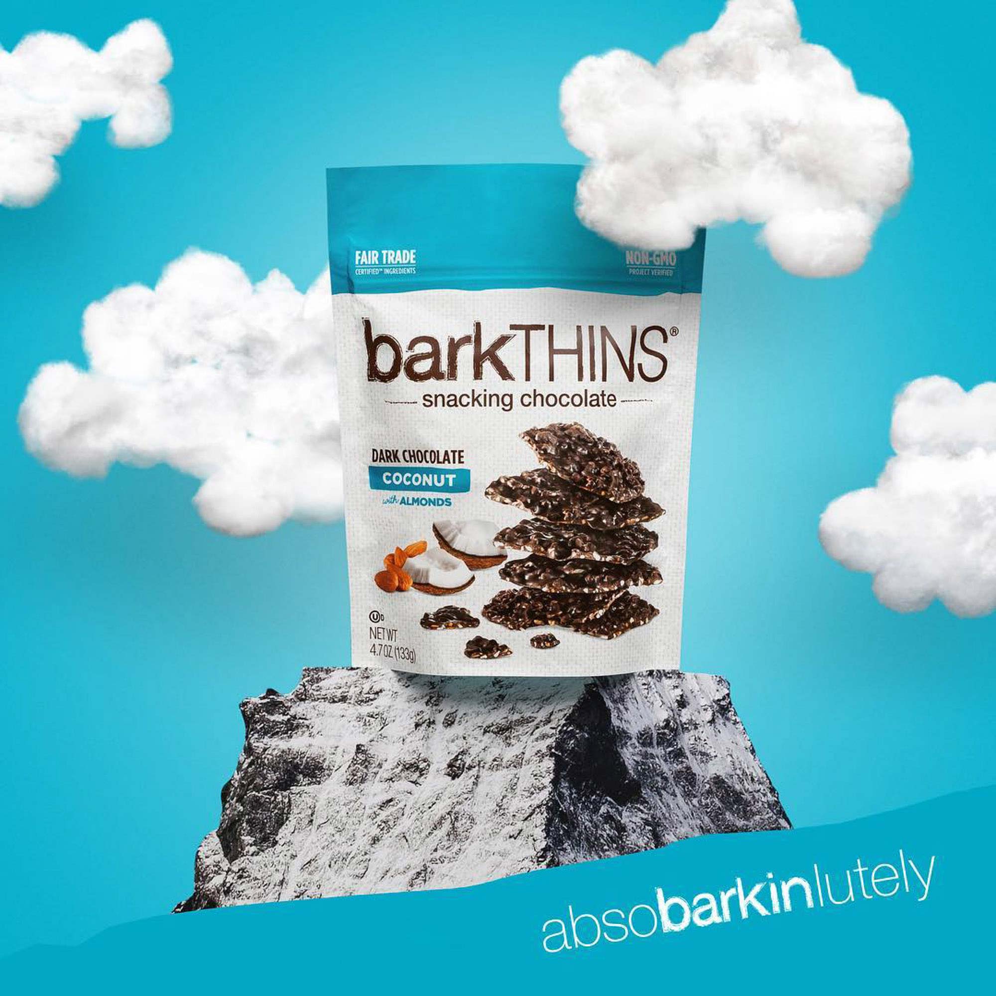 Product photograph of a barkTHINS Dark Chocolate Coconut bag perched on a rocky outcrop against a blue sky dotted with cotton-ball clouds, with the campaign tagline 'absobarkinlutely.'