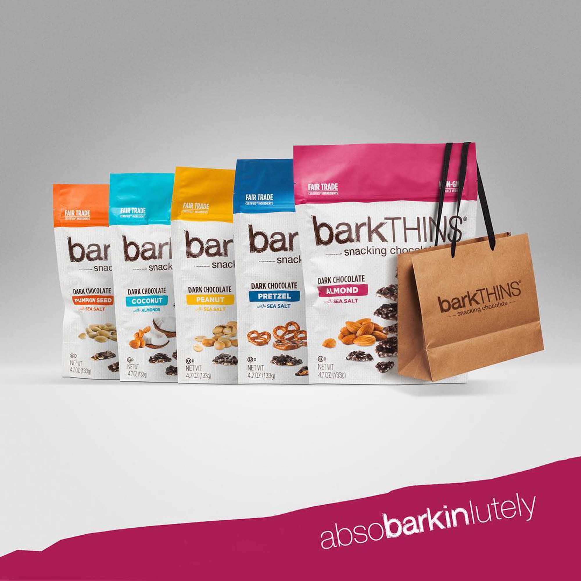 Five barkTHINS bags in a row — pumpkin seed, coconut, peanut, pretzel and almond flavors — alongside a branded paper shopping bag.