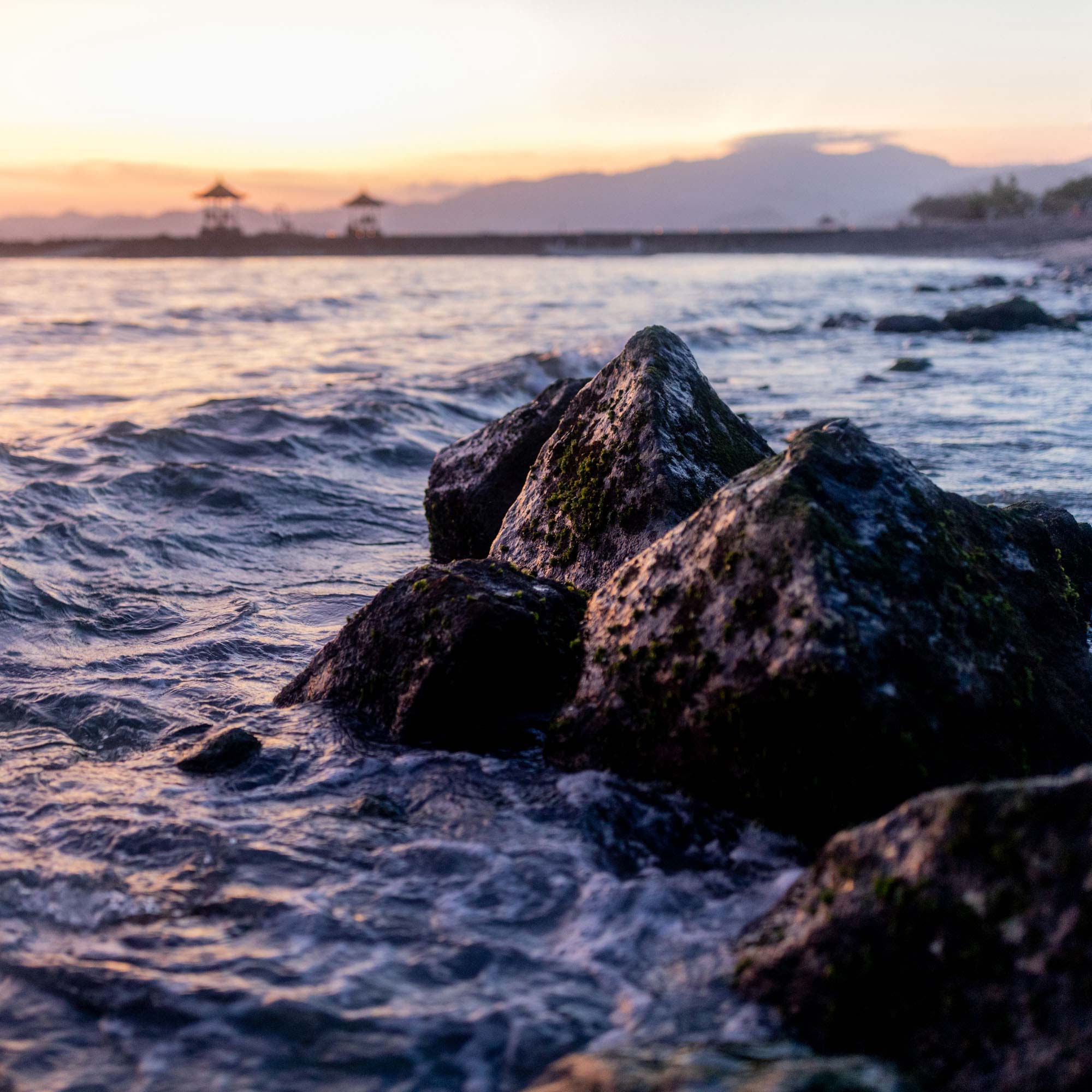 Waves break against moss-covered rocks at dusk near Candidasa, Bali, with beachside gazebos and mountain ridgelines silhouetted in the distance.