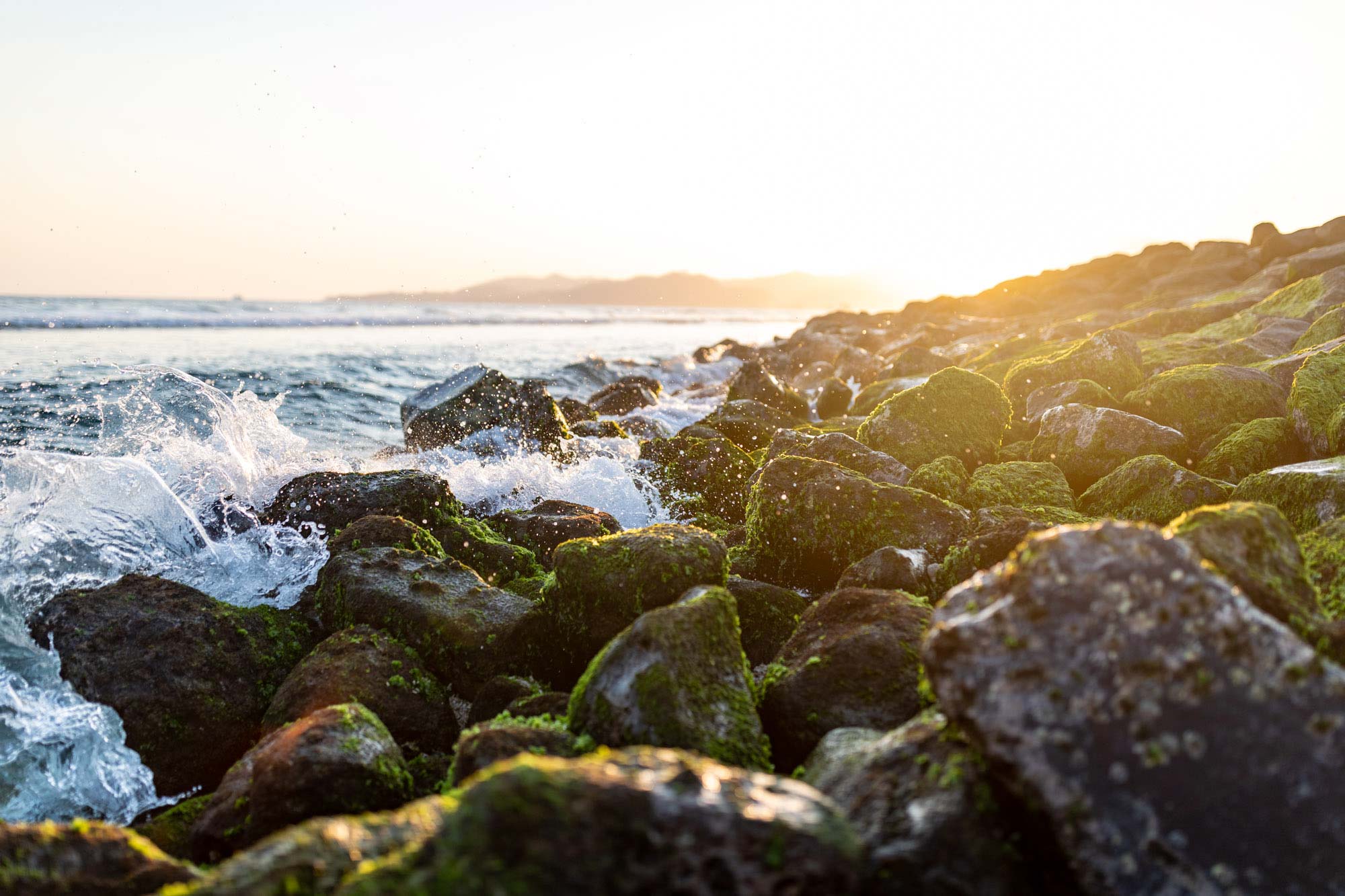 Golden sunset light catches moss-covered rocks and breaking surf along a Bali shoreline in Indonesia.