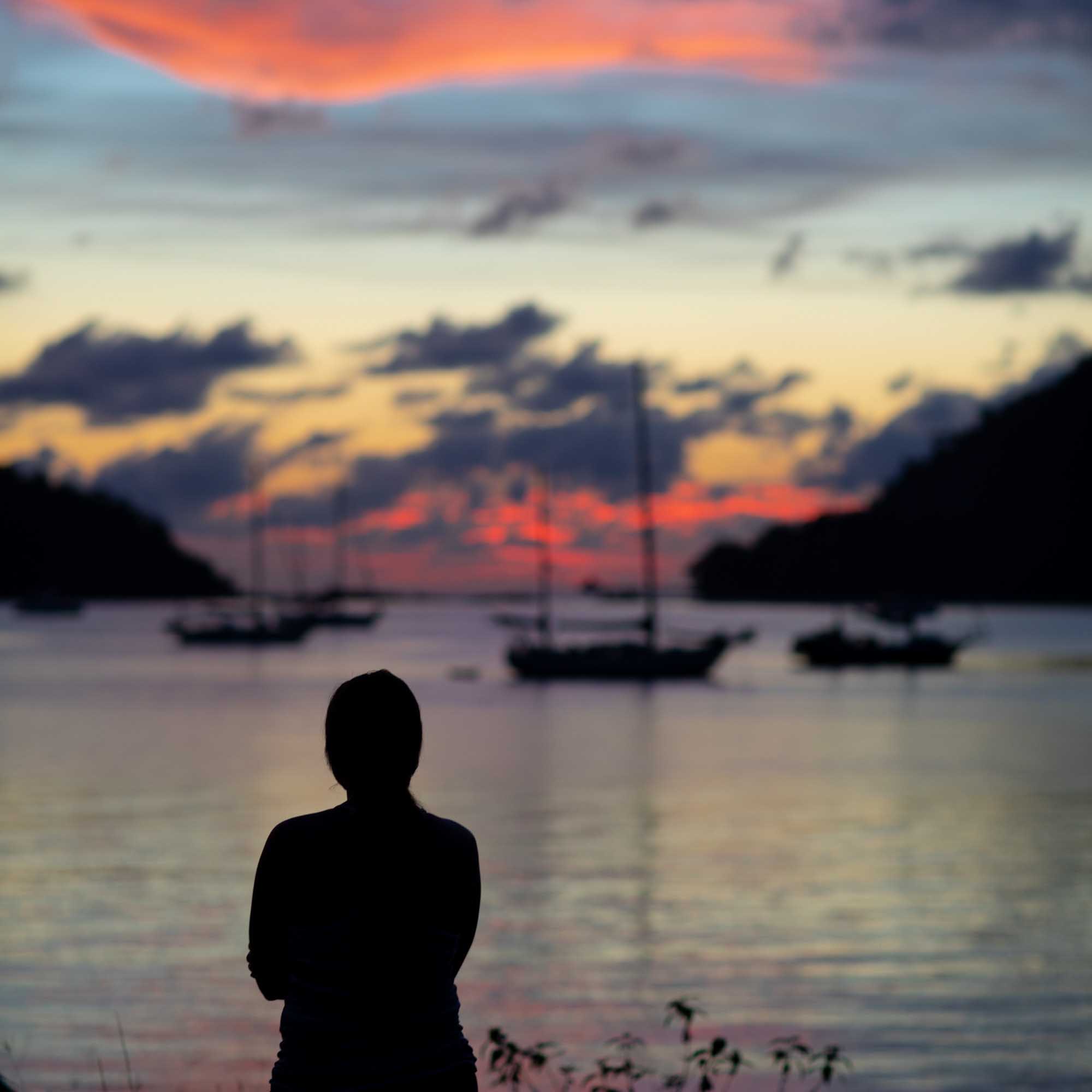 A silhouetted woman watches a pink-and-orange sunset over a harbor of anchored sailboats in Langkawi, Malaysia.