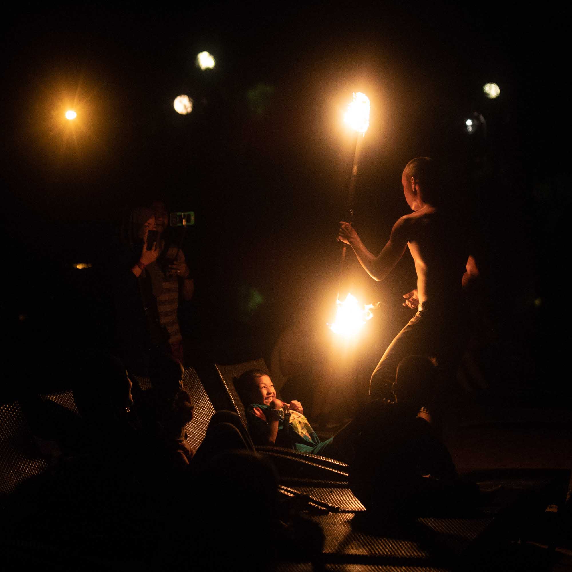 A child laughs on a reclining chair as a fire performer spins flaming staffs overhead at a nighttime show in Malaysia.