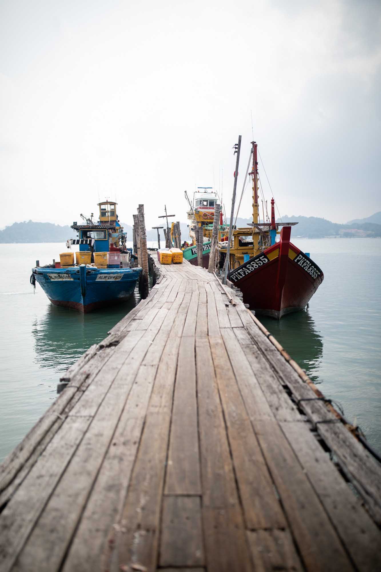 Wooden fishing boats moored along a weathered plank dock on an overcast day in Pulau Pangkor, Malaysia.