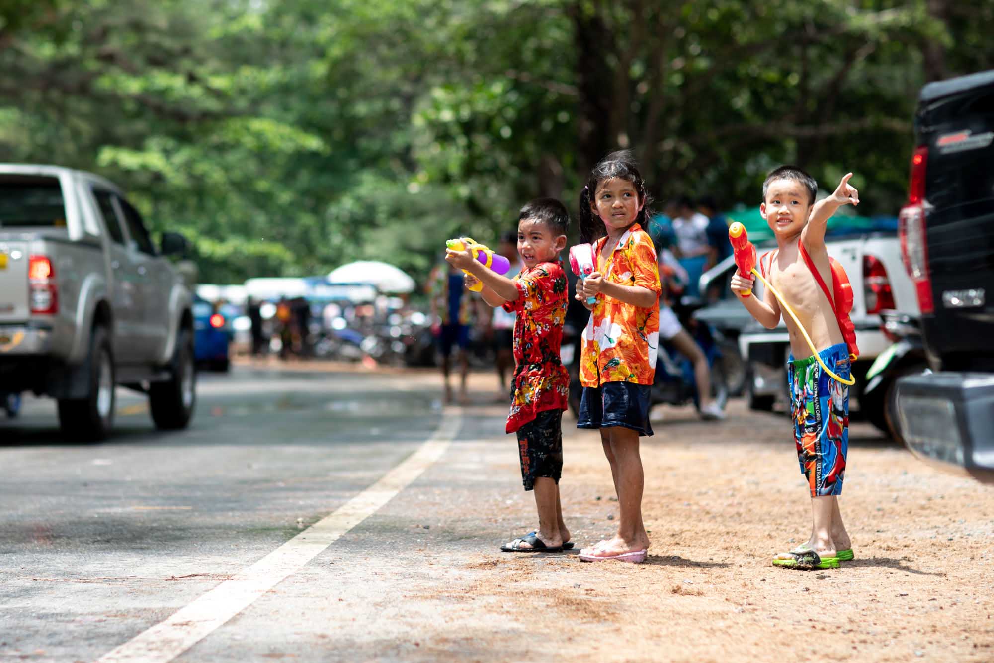 Three kids stand in a quiet Phuket street with super-soaker water guns, waiting for passing cars during the Songkran water festival.