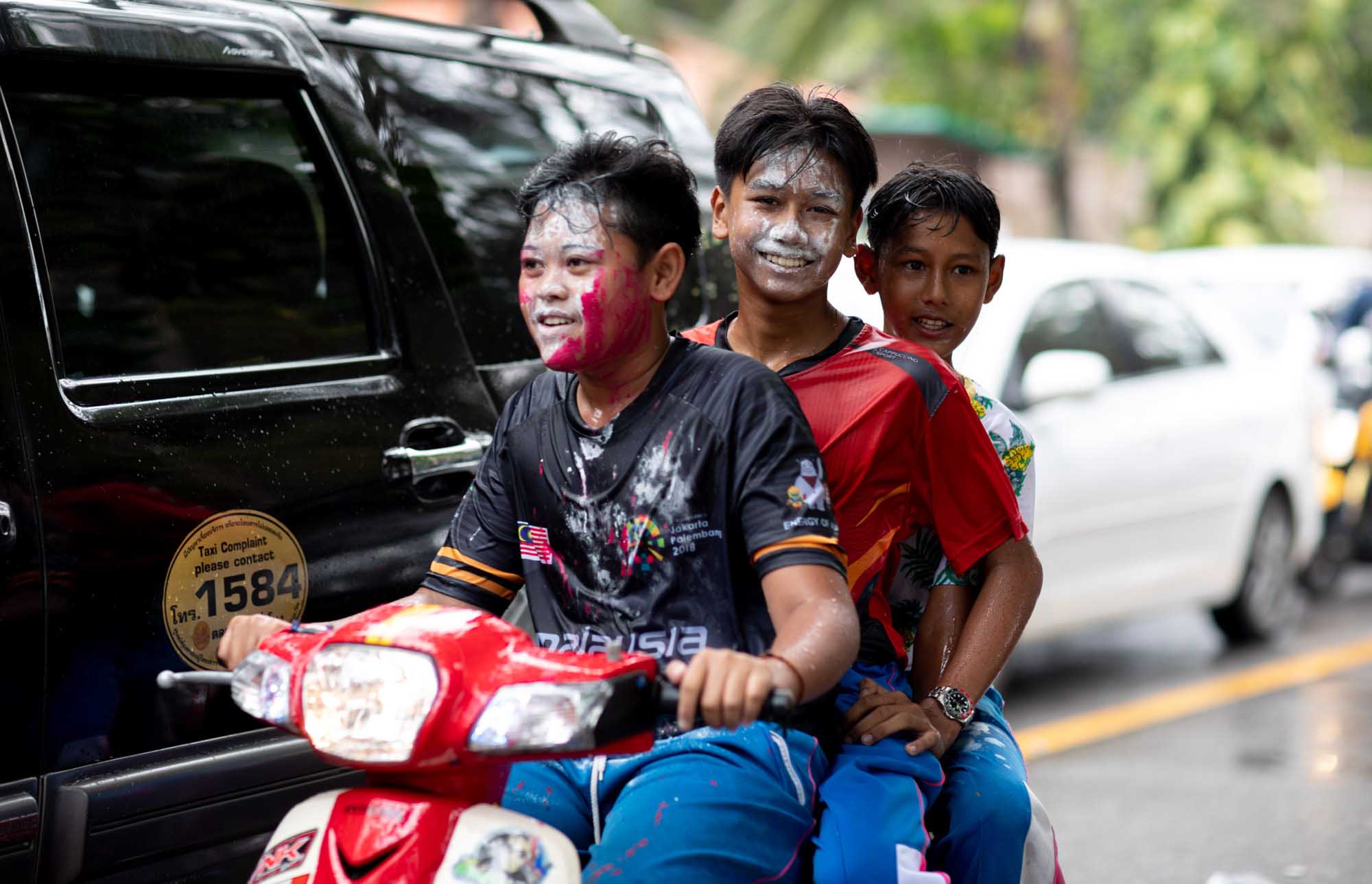 Three smiling boys squeezed onto a single red scooter, faces smeared with red and white powder, during the Songkran water festival in Phuket, Thailand.
