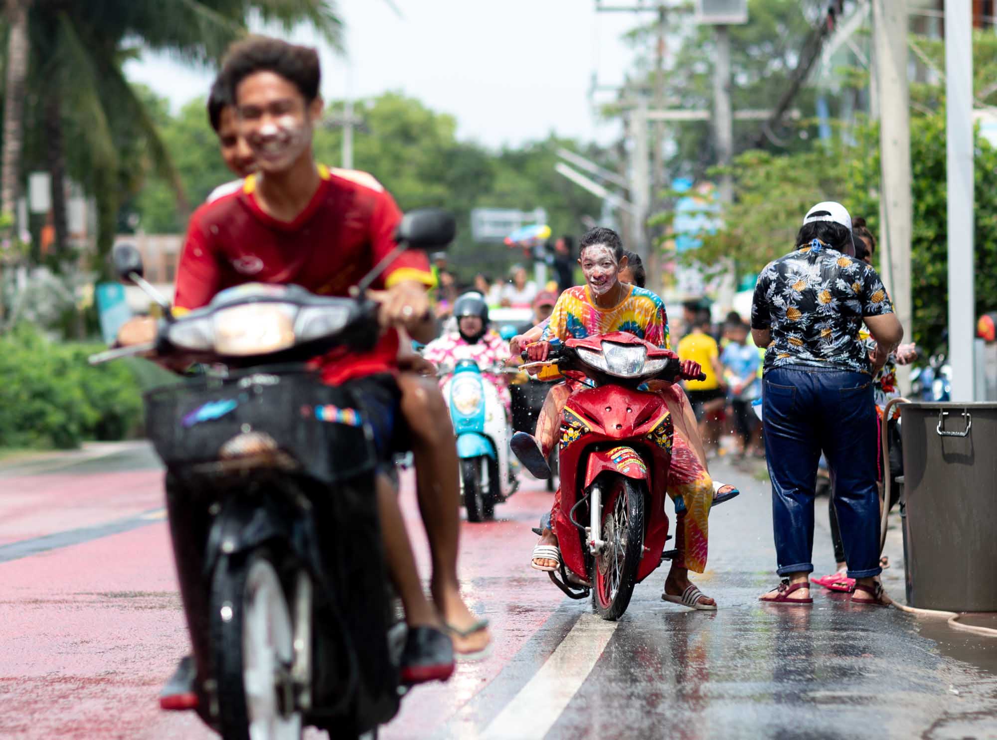 Teenagers on scooters ride through a Phuket street during the Songkran water festival, faces streaked with colored powder.