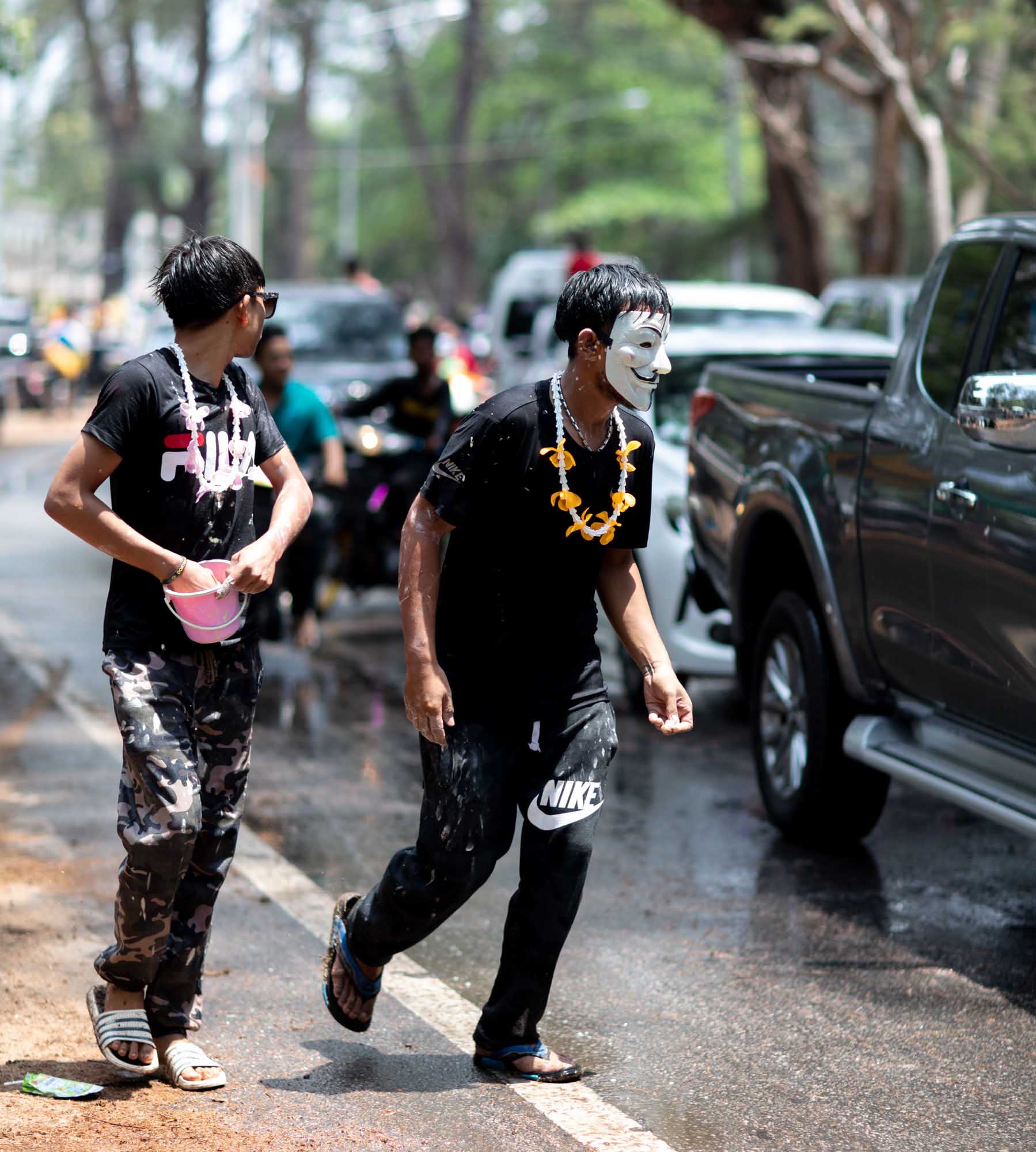 A young man in a Guy Fawkes mask jogs down the roadside during the Songkran water festival in Phuket, Thailand, a yellow flower lei around his neck.