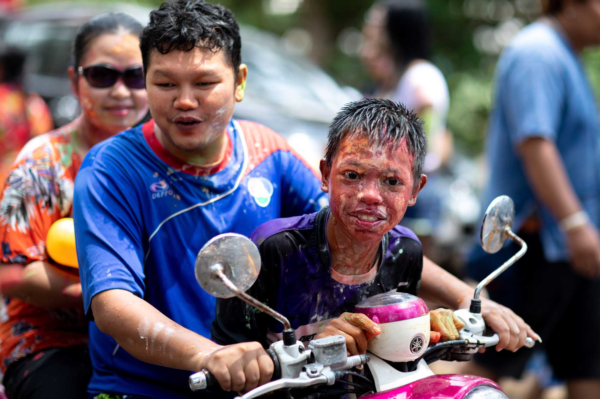 A boy rides pillion on a motorbike, face streaked with red powder, as a man in a blue sports jersey drives during the Songkran water festival in Phuket, Thailand.