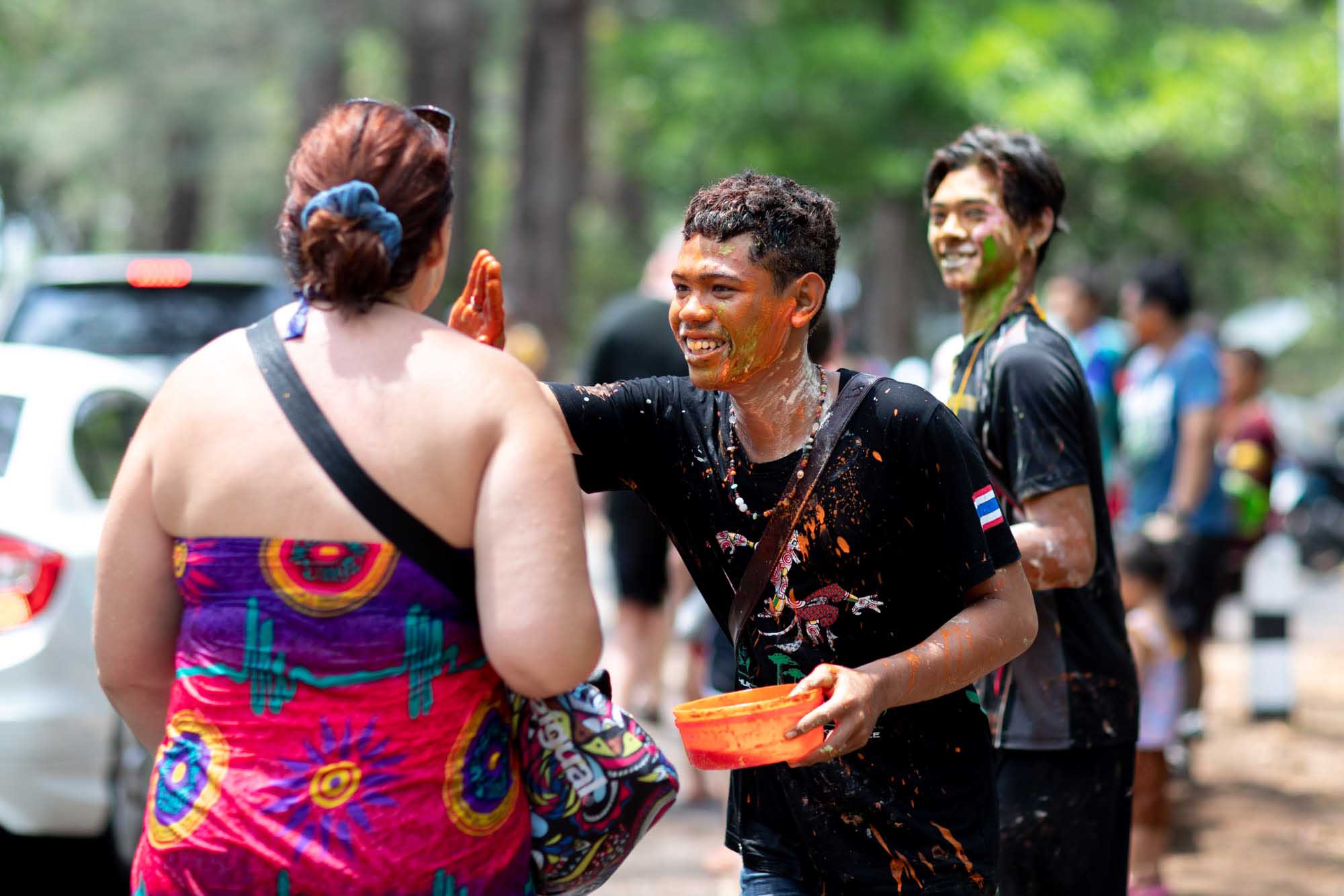 A woman in a colorful wrap dress raises her hand toward a man soaked in orange powder and holding a plastic bowl during the Songkran water festival in Phuket, Thailand.