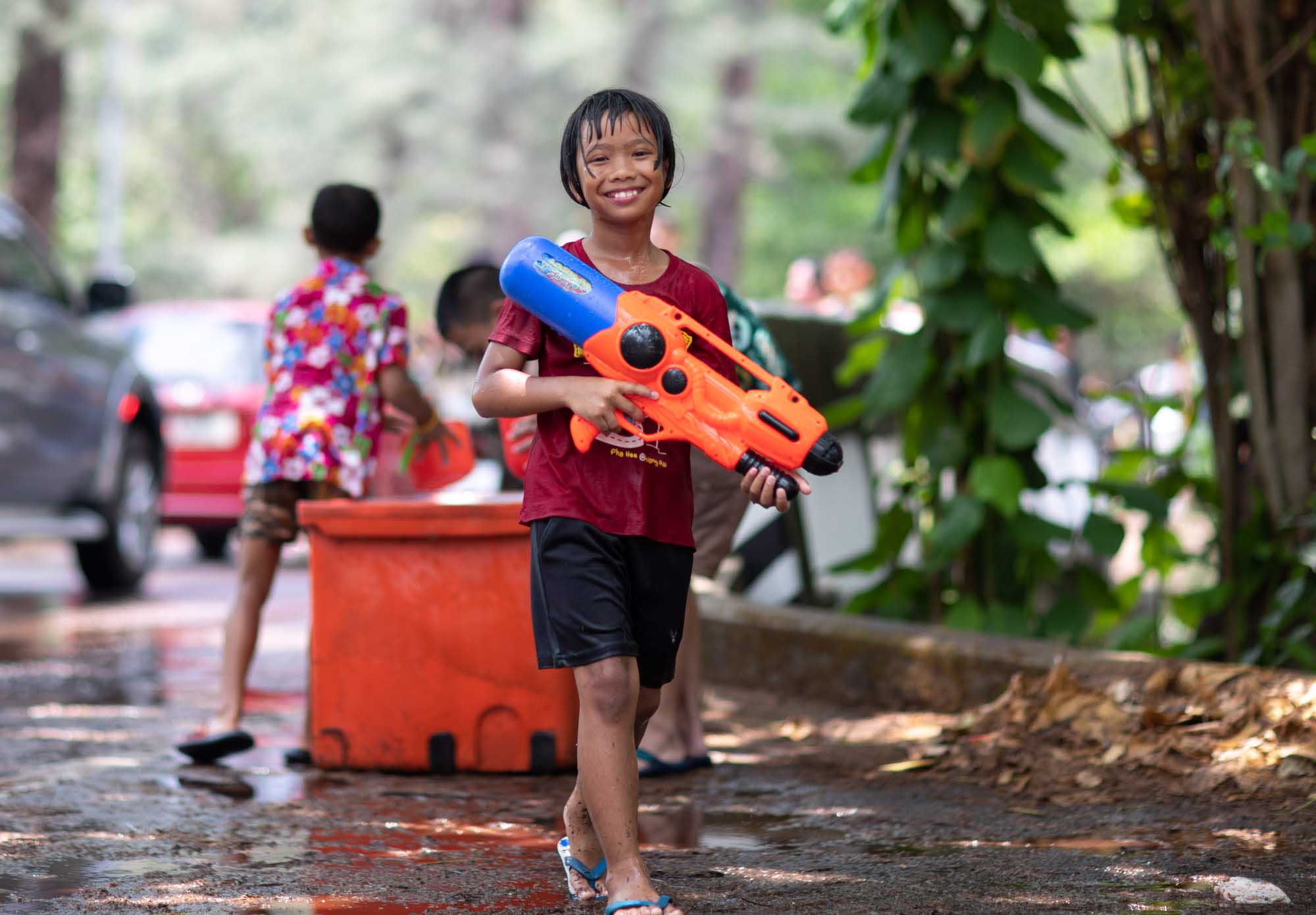 A soaking-wet boy in a red T-shirt smiles while hefting a large orange-and-blue water gun during the Songkran water festival in Phuket, Thailand.