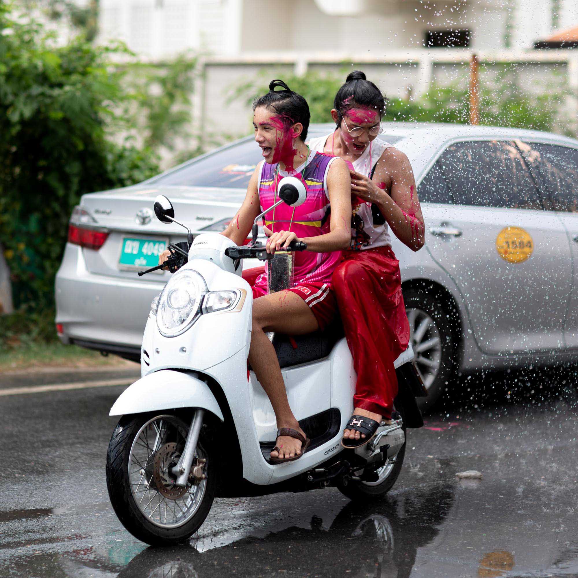 Two young women on a white scooter, faces smeared with red and pink powder, ride past a spray of water during the Songkran festival in Phuket, Thailand.