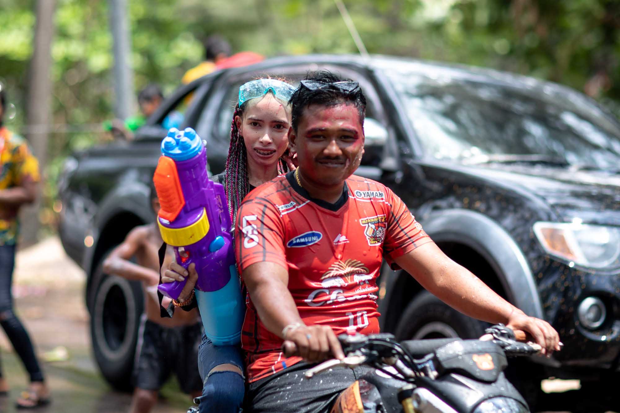 A couple rides a motorbike through a Phuket street during the Songkran water festival — he in an orange soccer jersey, she behind him with a purple water gun.