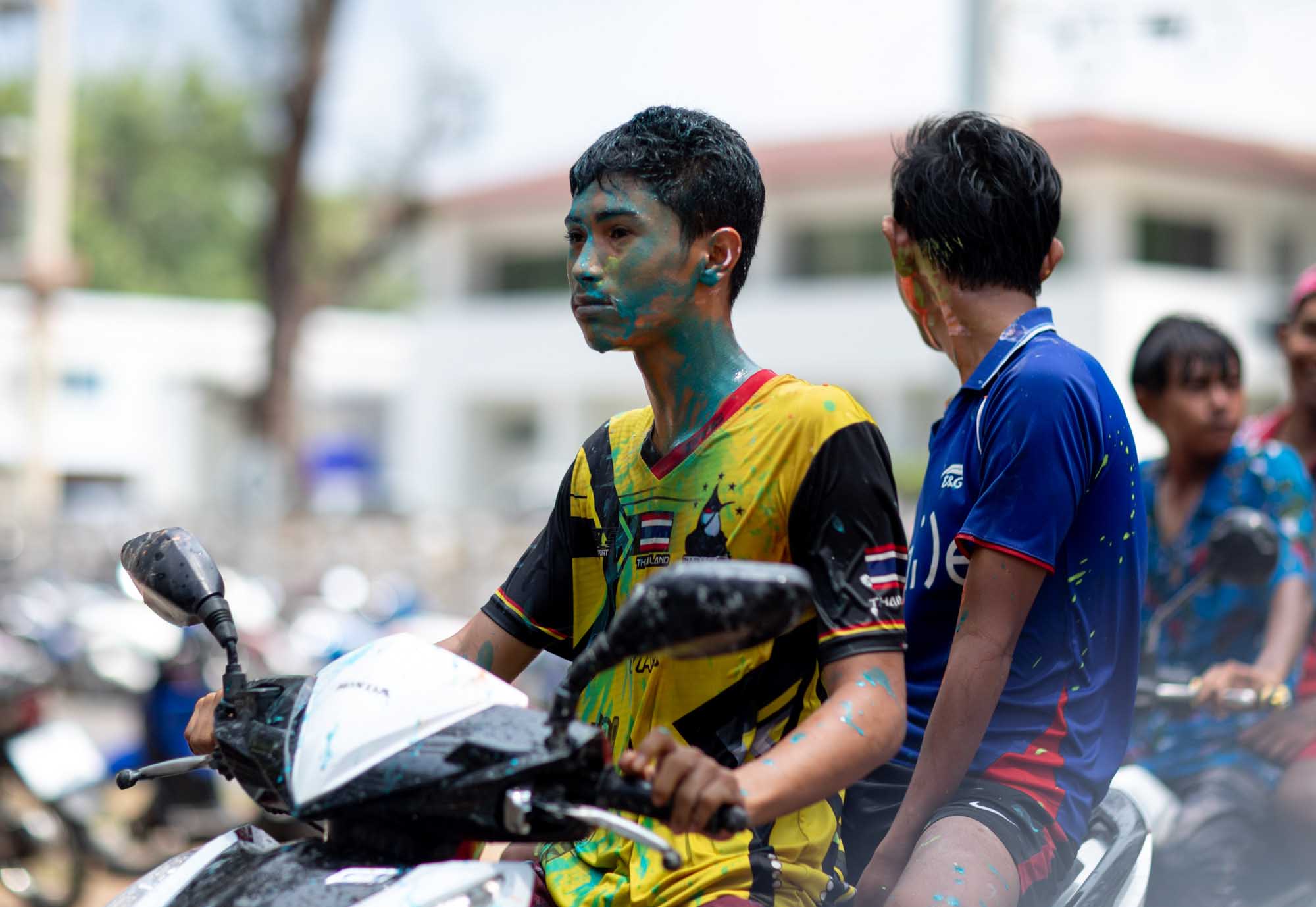 Two teenage boys on a motorbike, faces streaked with green and red powder, ride through a Phuket street during the Songkran water festival.