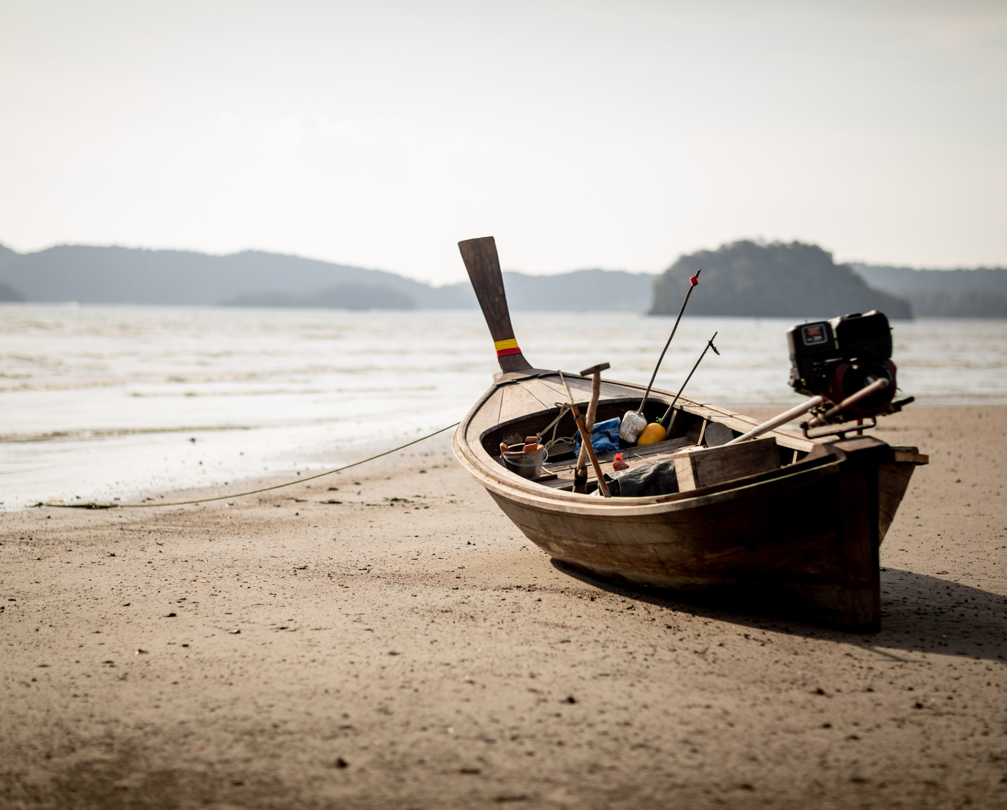 A wooden longtail boat beached on the sand at low tide with limestone karst islands in the distance — Ao Nang, Thailand.