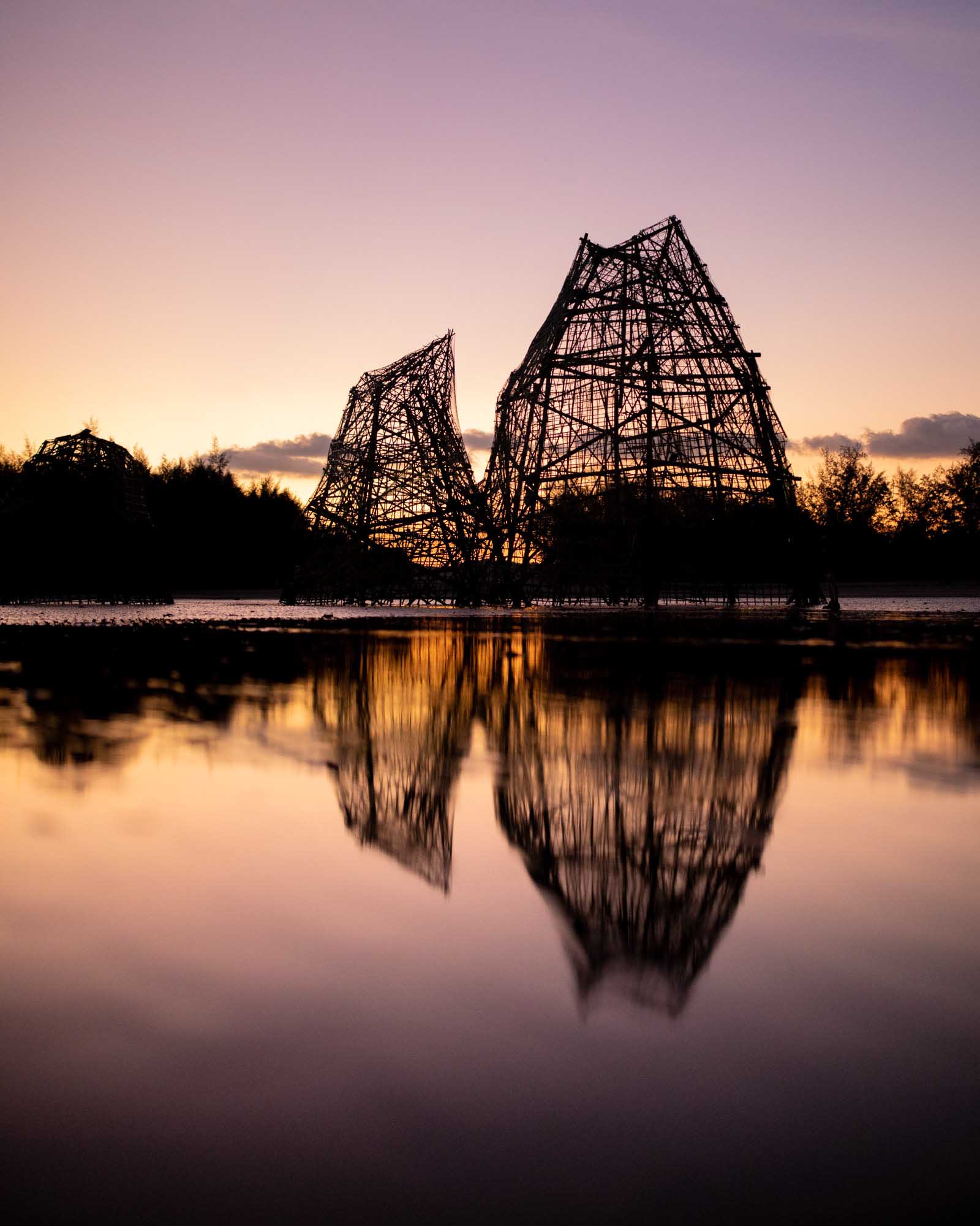 A large woven-wood sculpture silhouetted against a purple-orange sunset and reflected in still water — Ao Nang, Thailand.